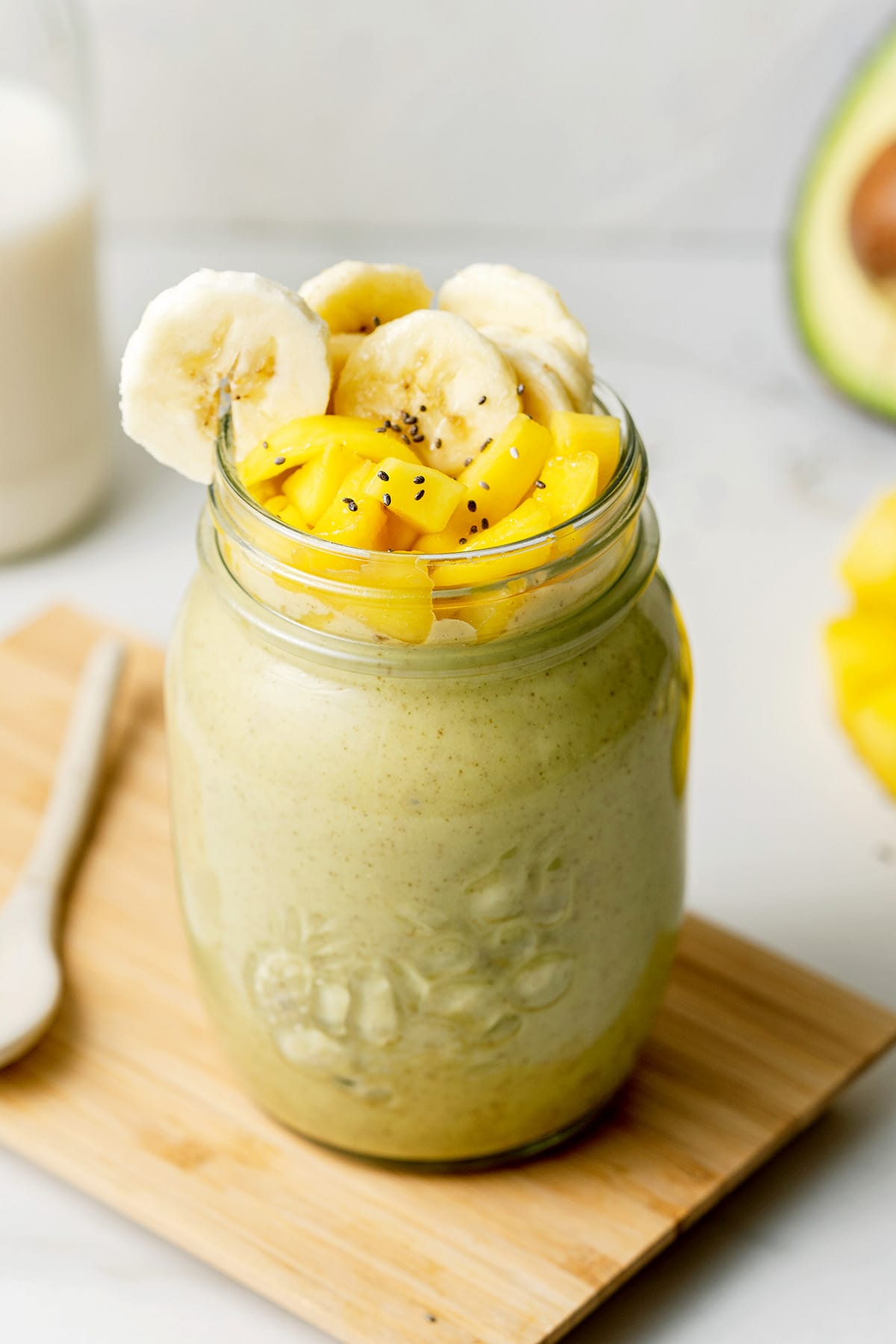 banana avocado smoothie in glass standing on wooden board. Smoothie topped with cut up fruit