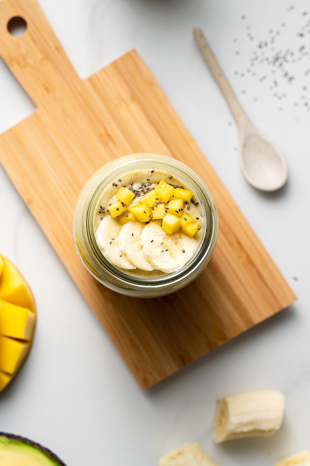 mango banana and avocado smoothie in jar on cutting board topped with banana slices and mango cubes