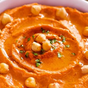 close up of roasted red pepper hummus in bowl with whole chickpeas, olive oil, and parsley topping