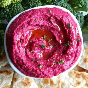 Pink beet hummus in white bowl, broccoli and pita bread triangles on the side
