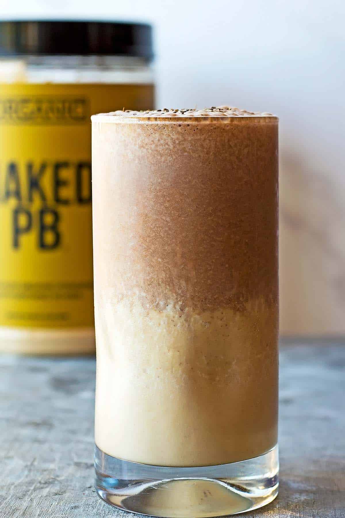 2 layered chocolate peanut butter protein shake in glass. peanut butter powder container in background