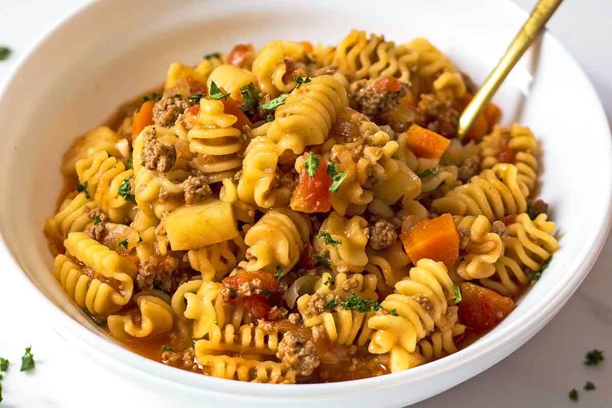 Ground Beef Stew Recipe with pasta in white bowl with golden utensil