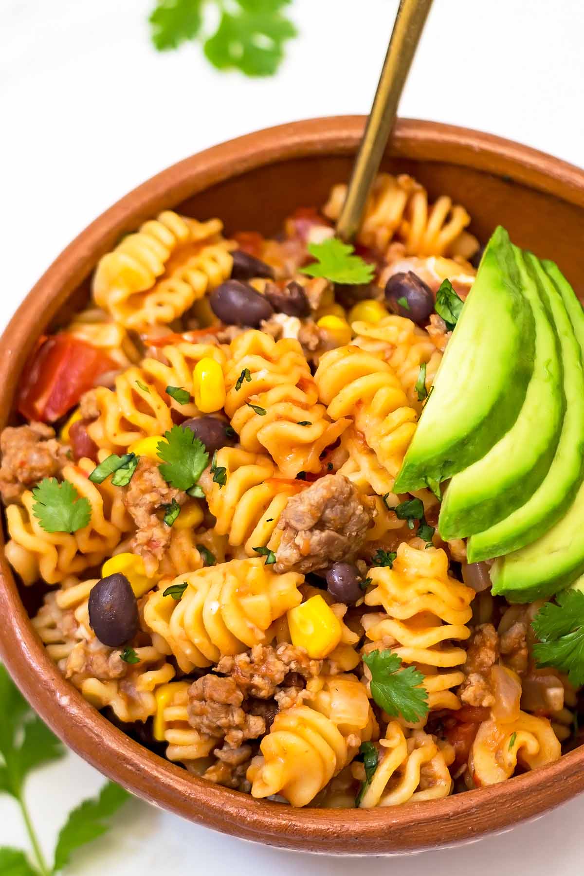 Taco pasta in Mexican clay bowl with avocado topping and golden fork inside