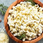 rosemary popcorn with garlic parmesan in wooden bowl, on the side: fresh rosemary sprig, garlic cloves, grated parmesan