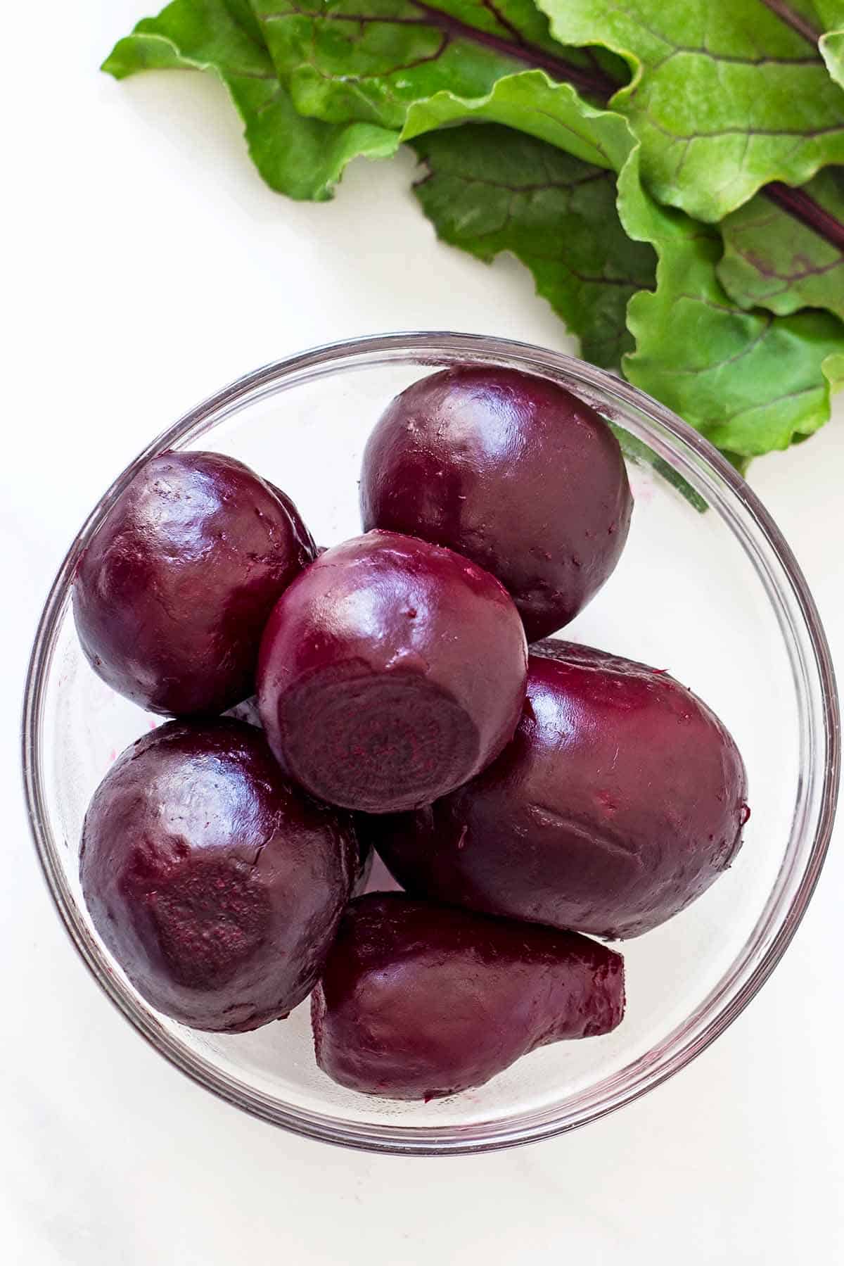 Peeled oven roasted beets in a glass bowl with beet leaves in background