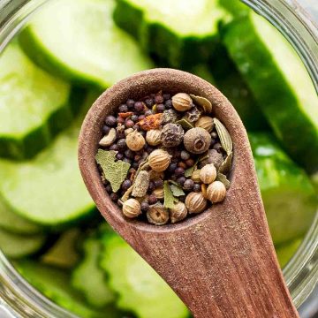 pickling spices on wooden spoon over glass jar of sliced cucumbers